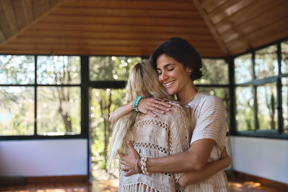 A blonde woman with her back to the camera hugging a brunette with a bob in a tranquil, peaceful room with large windows and wood beamed ceilings.