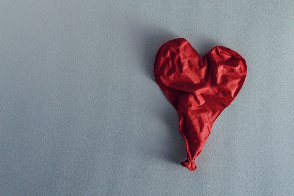 A deflated, red heart-shaped balloon on a gray background.  