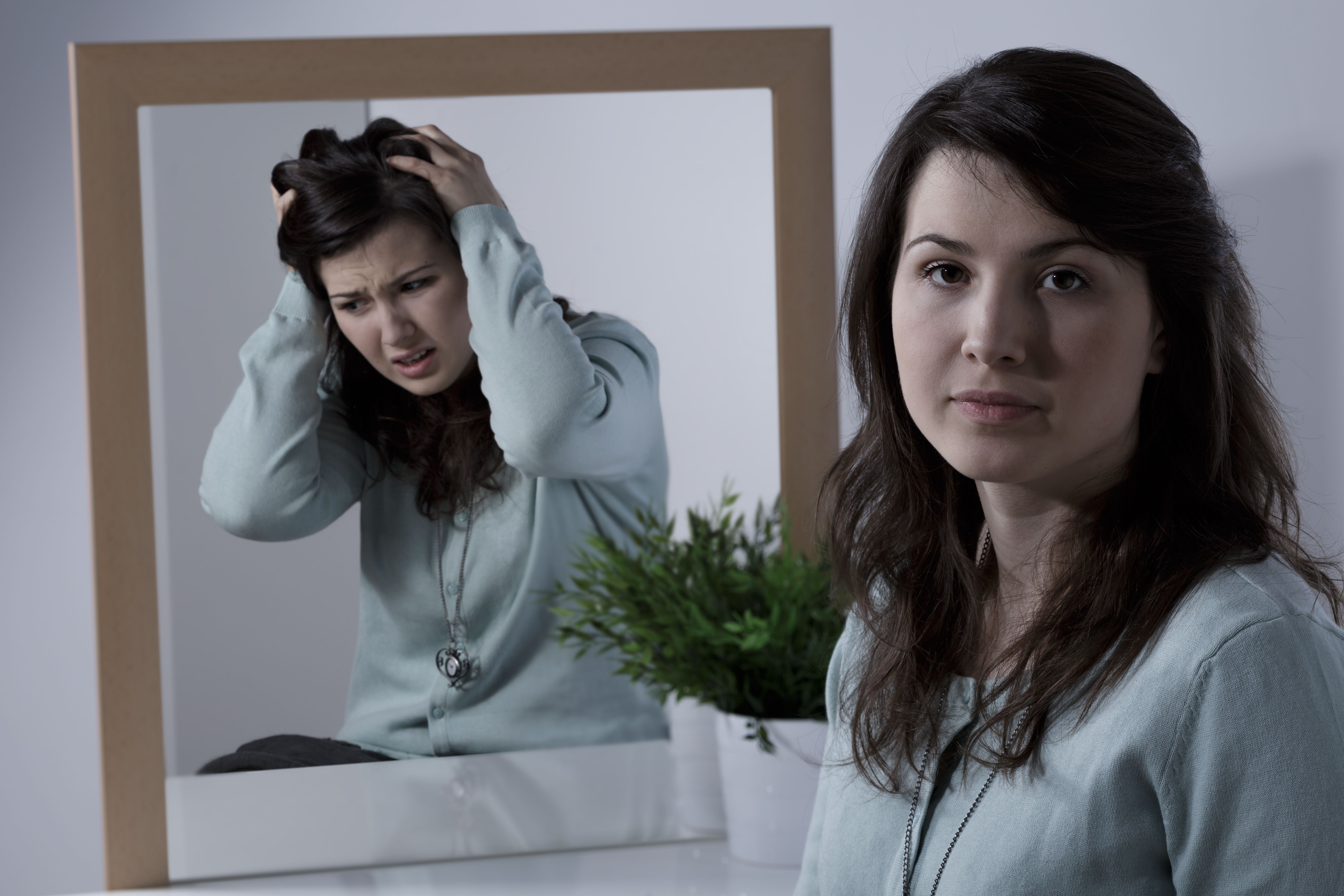 A young woman sits calmly in front of a mirror, while her reflection shows her distressed, pulling at her hair with a pained expression. The contrast highlights an inner struggle with emotions versus outward composure.